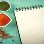 Drei Gewürze neben dem Gemüse (gelbe Paprika, rote Pepperoni, Knoblauchzehen), Kochlöffel und Notizblock
