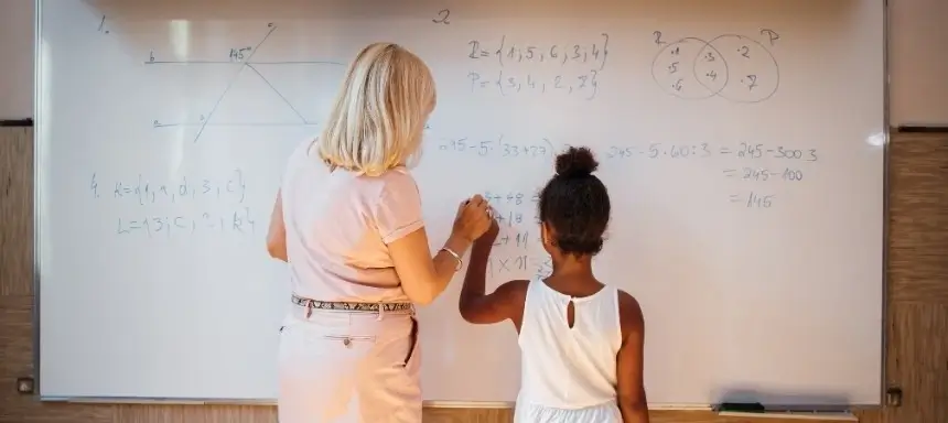 Ein Kind mit Dyskalkulie löst Matheaufgaben an einer Tafel und wird dabei von einer Lehrerin unterstützt.