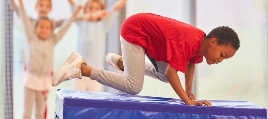 Kind springt seitlich über einen blauen Matratzenturm, im Hintergrund jubeln Kinder