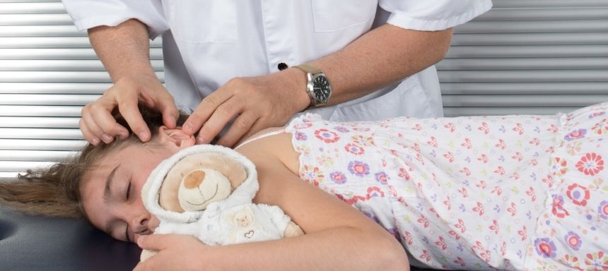 Arzt untersucht Mädchens Ohr, das auf den Bauch liegt mit Bär im Arm