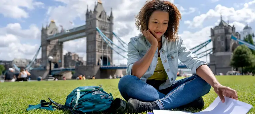 Eine Frau sitzt mit Unterlagen im Gras, im Hintergrund sieht man die Tower Bridge in London.